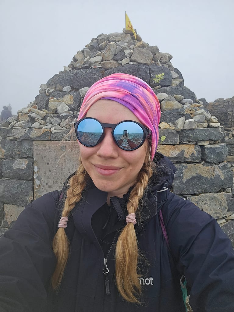 Una persona si scatta un selfie davanti a un monumento in pietra sulla cima del Ben Nevis. Indossa cappello e occhiali, con un cielo leggermente nuvoloso sullo sfondo.