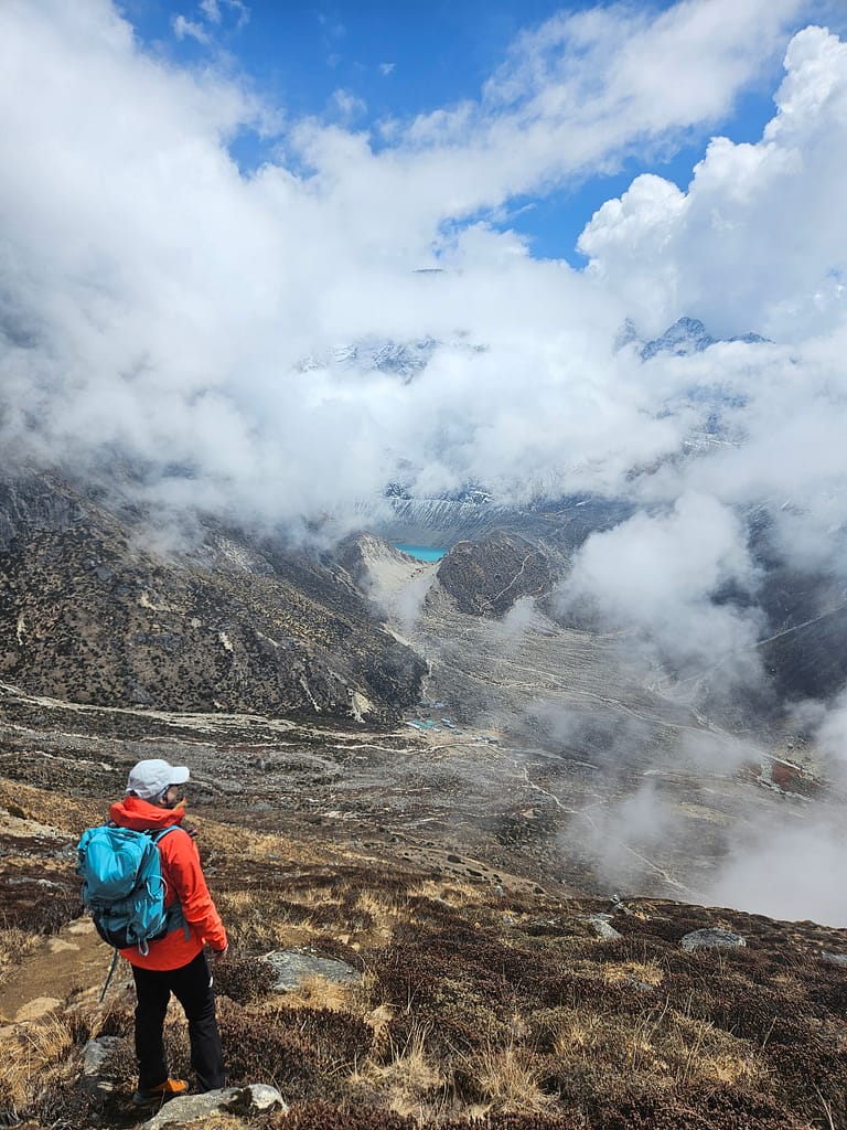 Yürüyüş yapan bir dağcı, bulutların arasında mavi bir göl ve yüksek dağ manzarasına bakıyor.
