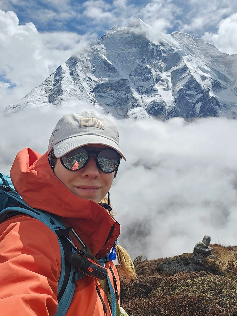 Dağ manzarasına karşı öz çekim yapan bir dağcı, turuncu bir mont giymekte ve güneş gözlüğü takmaktadır. Arkada, karla kaplı yüksek dağlar ve bulutlar yer almakta.
