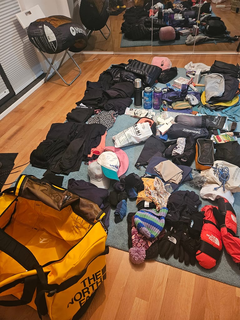 Tırmanış için hazırlanan dağcılık ekipmanları ve giysileri bir odada düzene konmuş. Sarı bir çanta açık halde ve çeşitli dağcılık aksesuarları, giysiler, şapkalar, eldivenler, su şişeleri ve diğer malzemeler yerde serilmiş.