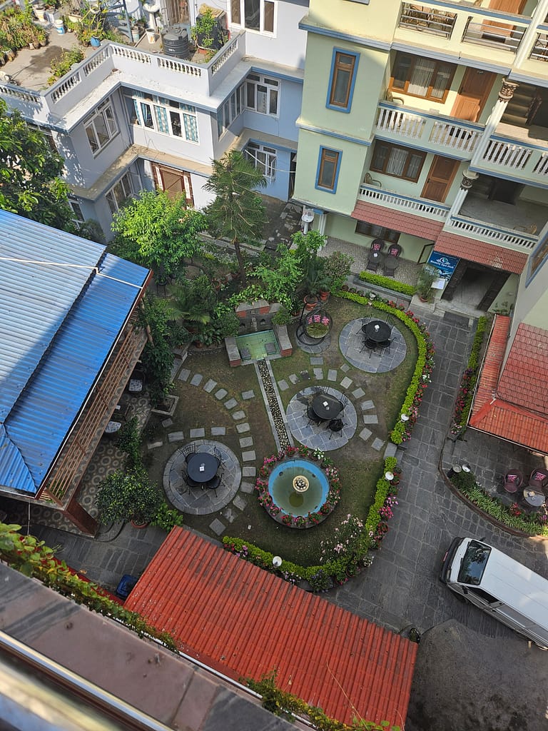 Bir otelin avlusunun yukarıdan görünümü, içerisinde yeşil bitkiler ve çiçeklerle düzenlenmiş dinlenme alanları bulunan bir bahçe.