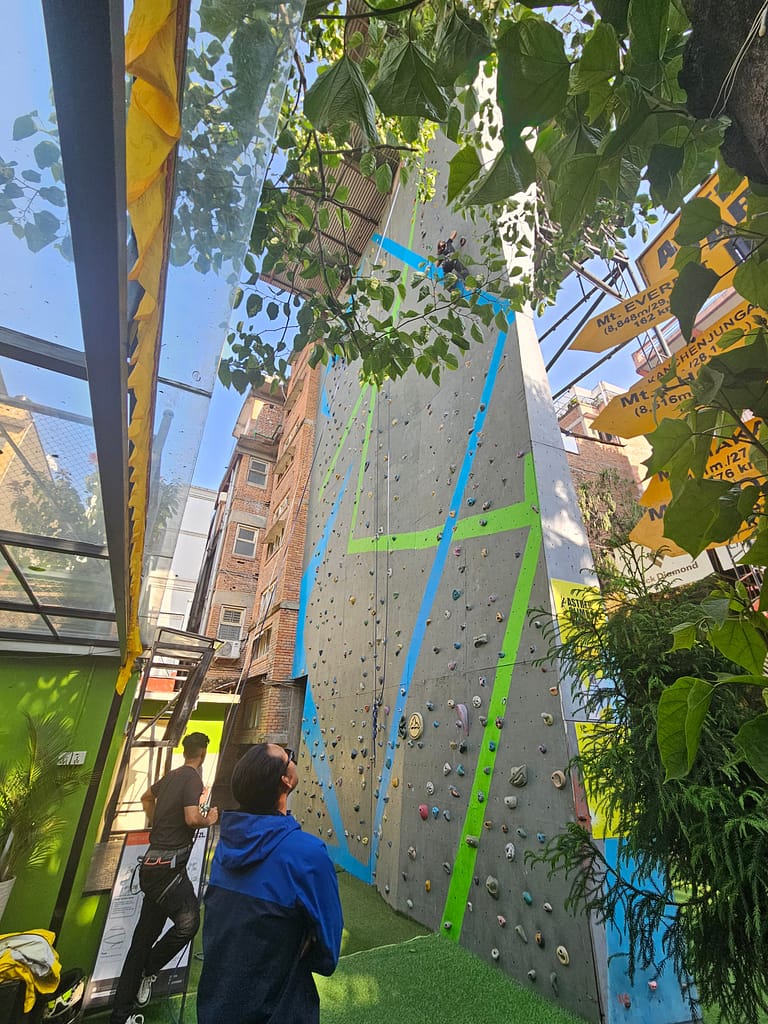 Bir tırmanış duvarının önünde, tırmanış yapan kişiyi izleyen iki kişi. Tırmanış duvarı yeşil ve mavi çizgilerle işaretlenmiş, çevresinde bitkiler ve binalar var.