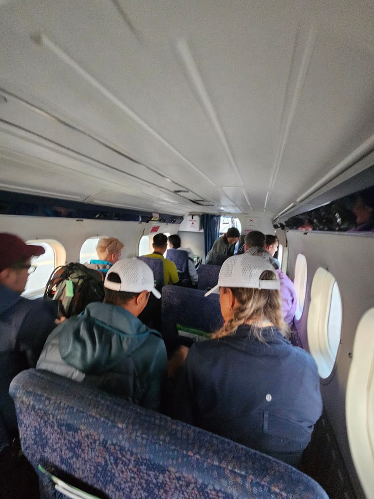 İçerisi bir uçağın kabinini gösteren fotoğraf. Uçak, yolcularla dolu; bazı yolcular kafalarını aşağıya eğmişken, bazıları kıyafetlerinin üzerine beyaz şapka giymiş. Uçak koltuklarının yer aldığı alan görünmekte.