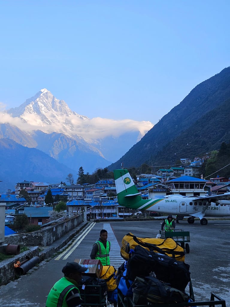 Lukla Havalimanı'na iniş sırasında, dağ manzarası ve mavi çatılarıyla köy arka planda yer alıyor. Aşağıda bagajlarını taşıyan ekip üyeleri görünüyor ve bir uçak beklemede.