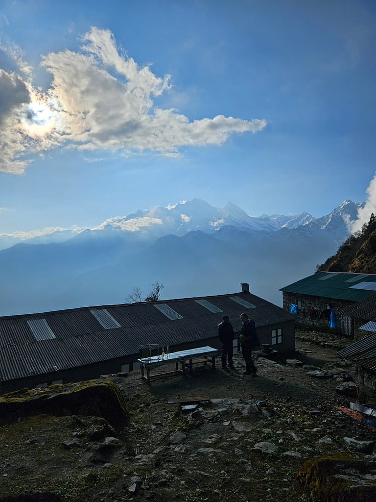 Yüksek dağlarla çevrili bir bölgedeki tea house'un önünde iki kişi durmakta. Gökyüzünde bulutlar, dağların zirvelerinin arkasında görünmekte.