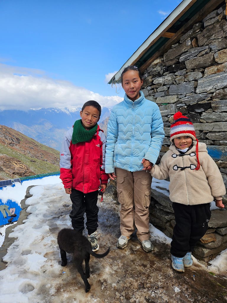 İki çocuk ve bir genç kız, Himalayalar'da bir dağ evinin önünde duruyor. Arka planda dağ manzarası görünmekte. Çocuklardan biri yeşil bir atkı takarken, diğerinin kafasında renkli bir şapka var. Kız, mavi bir mont giymekte ve bir çocuğun elini tutuyor.