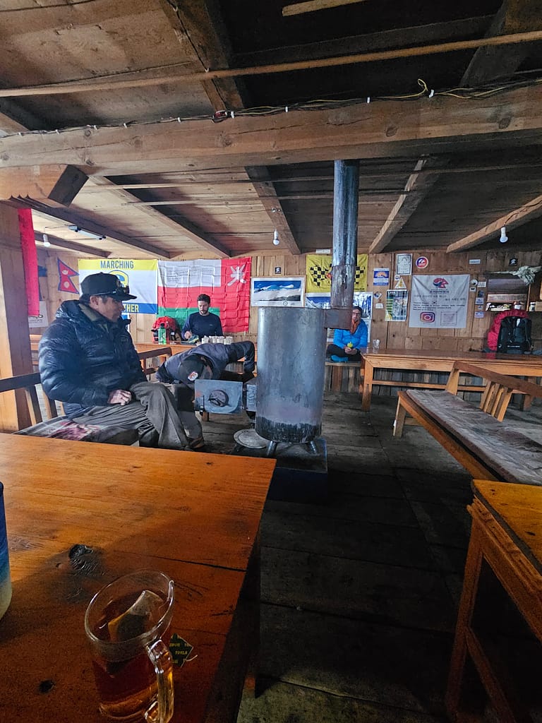 Bir dağcı grubunun oturup dinlendiği bir dağ evinin içi, odanın ortasında bir soba, duvarlarda çeşitli bayraklar ve posterler var. Ahşap masalar ve sandalyeler, sıcak bir atmosfer oluşturmakta.