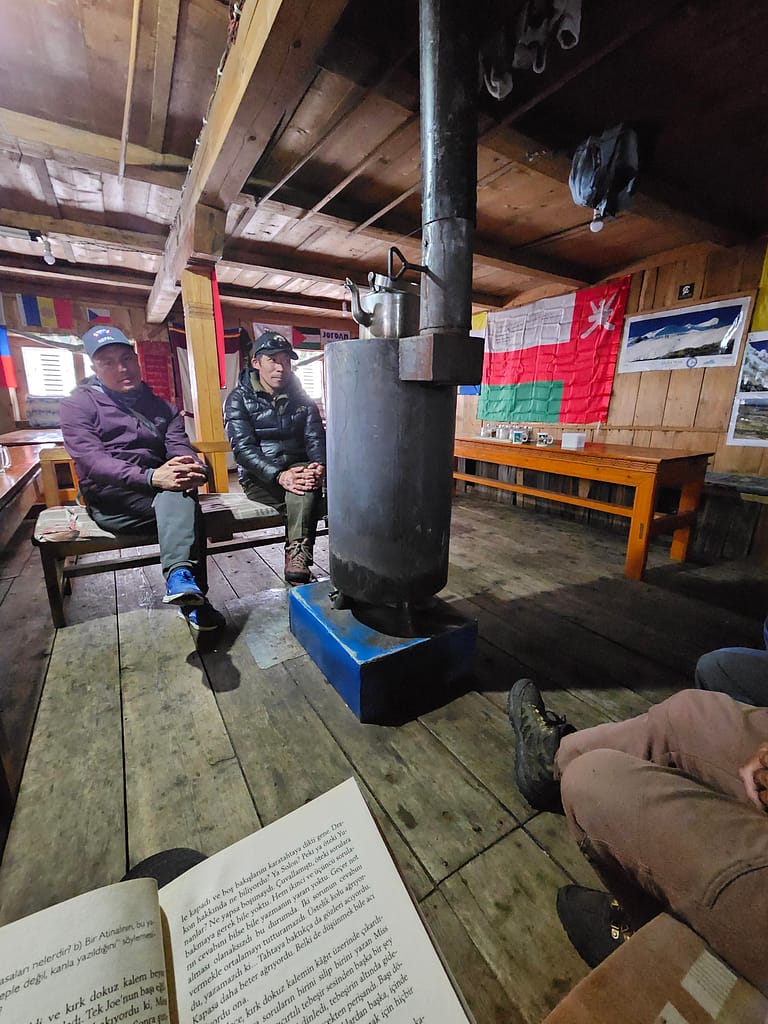 Bir grup insan, ahşap bir tesiste oturuyor. Ortada bir soba var ve odanın duvarlarında çeşitli bayraklar asılı. Sol köşede bir kitap açık bir şekilde yer almakta.