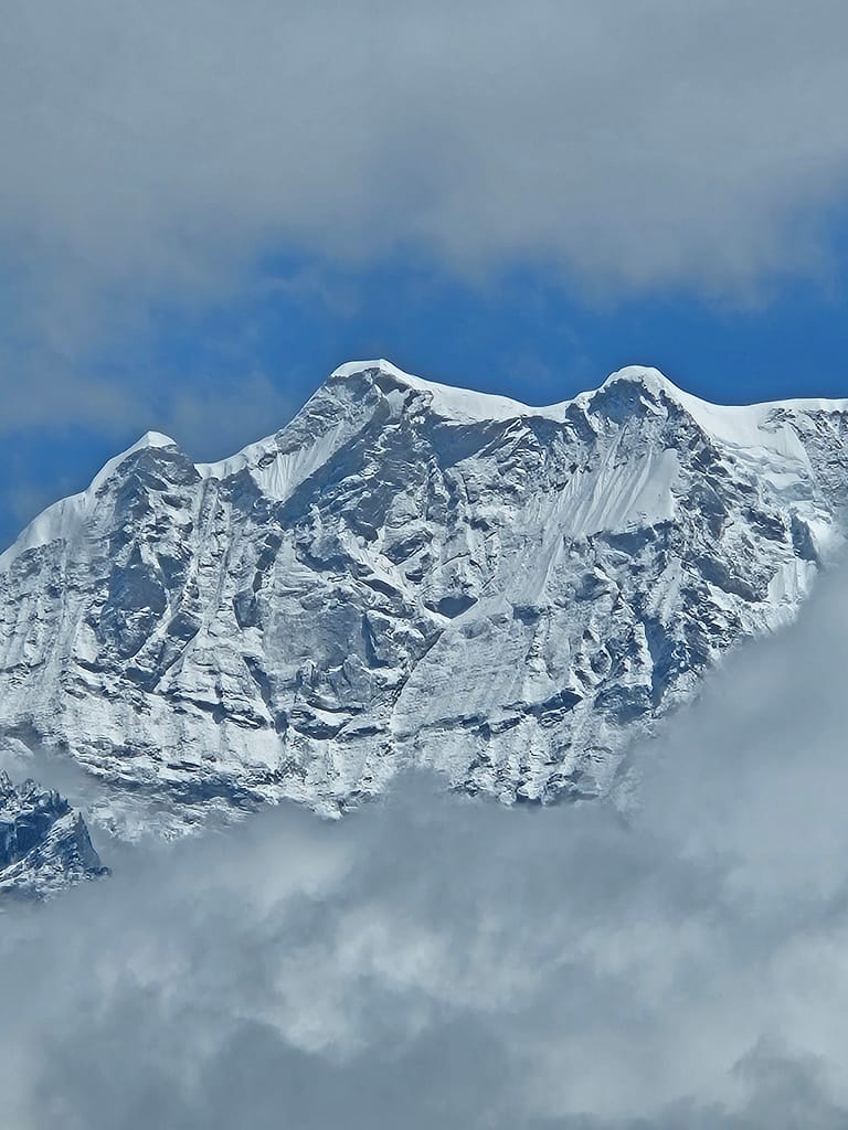 Yüksek dağ zirvesi, karla kaplı ve bulutlarla çevrili, mavi gökyüzü arka planda.