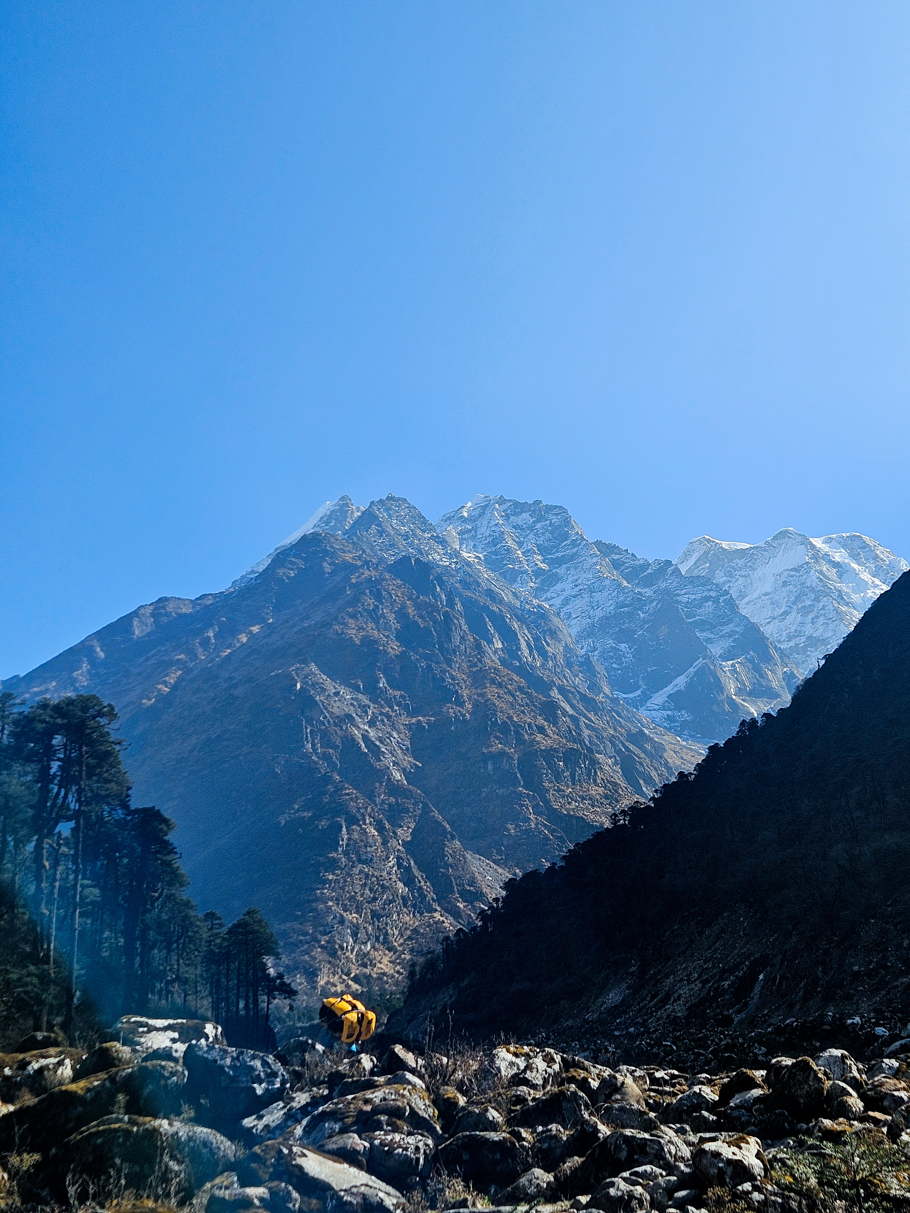 Dağ manzarası eşliğinde yürüyüş yapan bir kişi, çantasıyla kayalık bir patikadan yükseliyor. Arka planda yüksek, karla kaplı dağlar ve mavi gökyüzü bulunuyor.