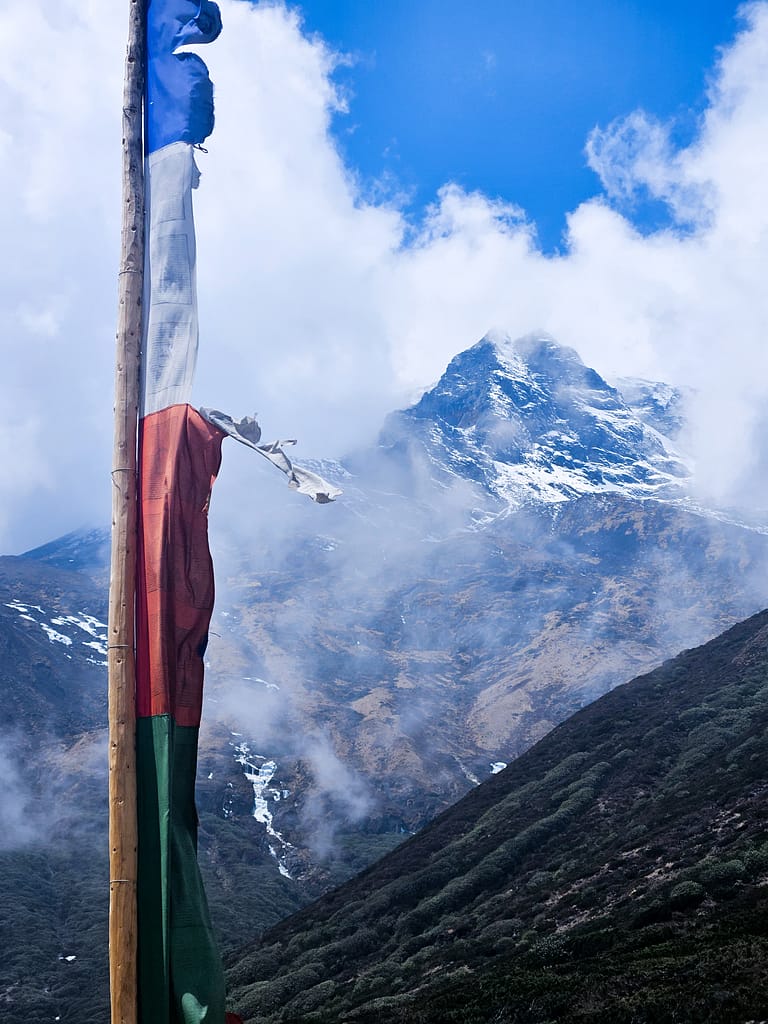 Rüzgarda dalgalanan dua bayrağı ön planda, arka planda karla kaplı dağ zirvesi ve bulutlu bir gökyüzü görülüyor.