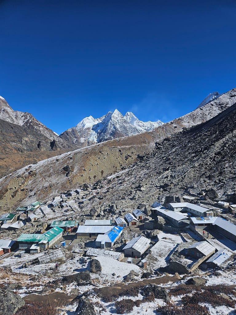 Yüksek dağların eteklerinde yer alan kış koşullarında bir köy manzarası. Karla kaplı zemin üzerinde metal çatıya sahip yapılar görünmekte. Arka planda karla kaplı zirveler yer alıyor ve gökyüzü açık mavi.