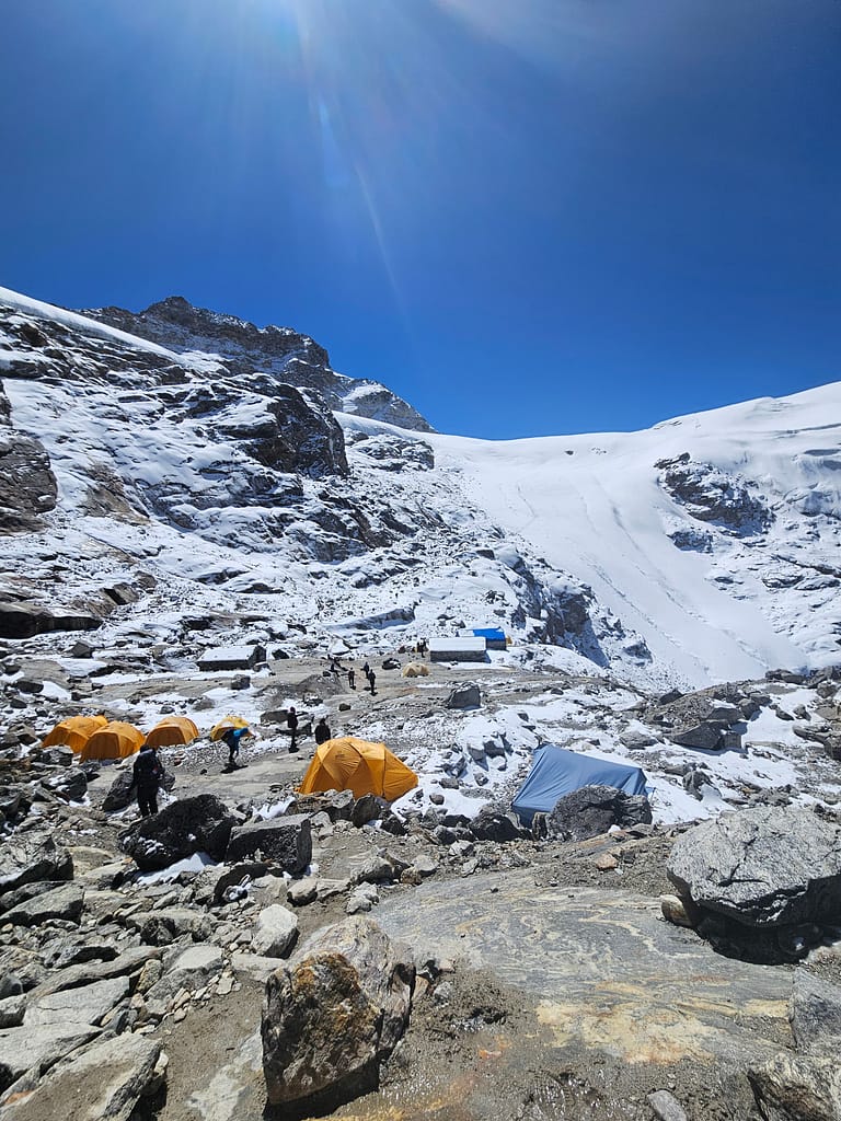 Karlarla kaplı dağlık bir alanda, farklı renklerde çadırların bulunduğu bir kamp alanı. Düşük bir güneş ışığı altında, mavi gökyüzü ve uzak dağ manzarası.