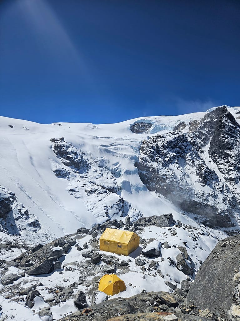 Yüksek irtifada kar ve buzul manzarasının bulunduğu bir alanda kurulu sarı çadırlar ve kayalıklar.