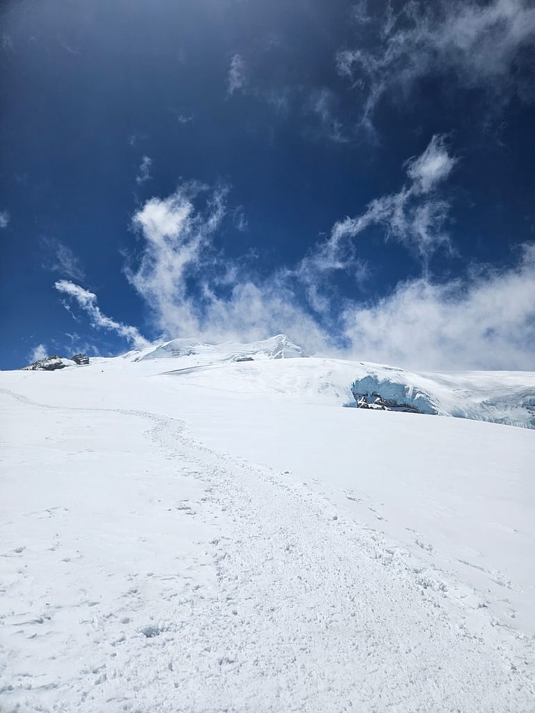 Buzul ve kar kaplı dağ manzarası, mavi gökyüzü ve beyaz bulutlar ile yüksek irtifada bir yürüyüş rotası.
