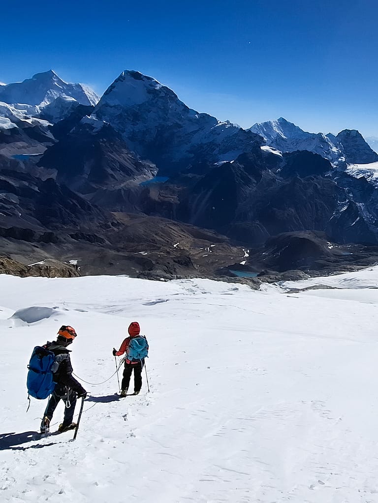 İki dağcı, kalın kar örtüsü üzerinde ilerlerken yüksek dağ manzarası arka planda.