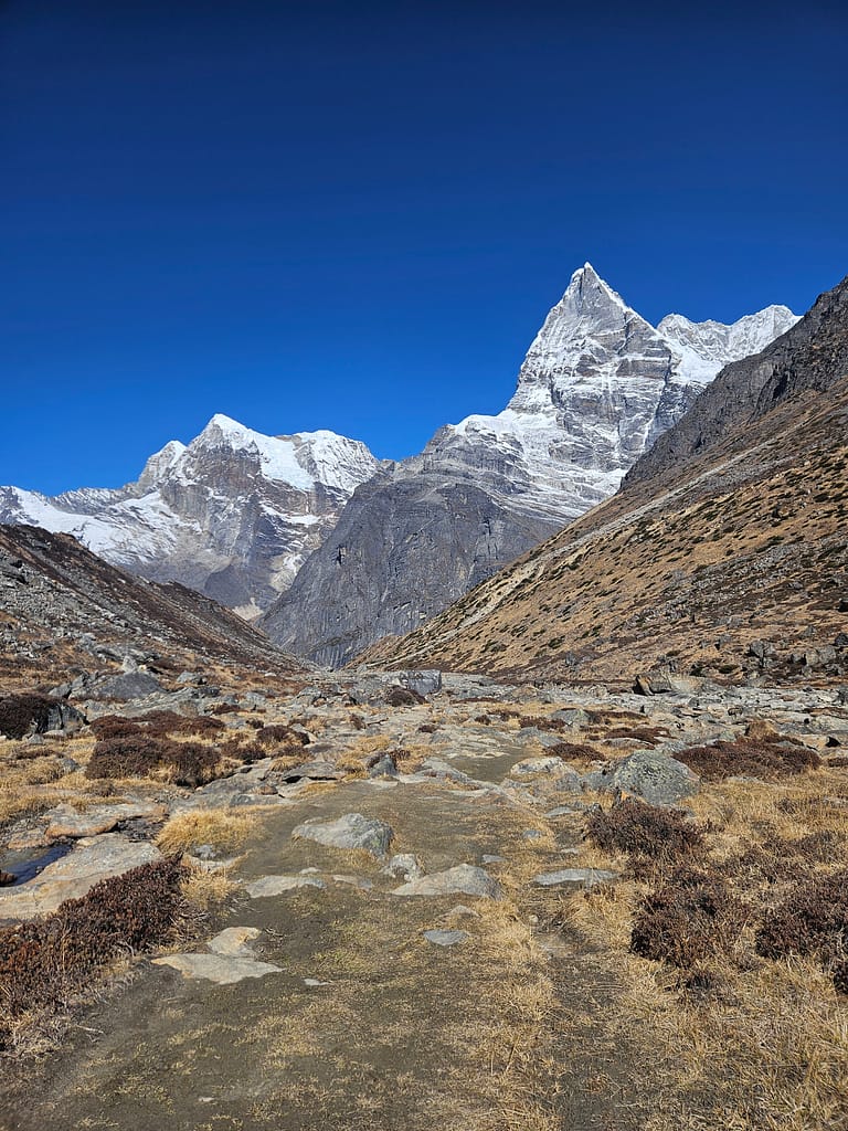 Geniş bir dağ vadisi manzarası, arka planda karla kaplı yüksek zirveler ve temiz mavi gökyüzü ile doğal bir yürüyüş yolu.