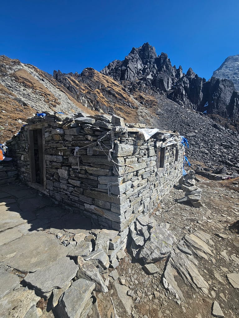 Dağlık bir alanda, taşlardan yapılmış bir yapının dış görünümü. Arkada yüksek dağlar ve mavi gökyüzü görünüyor.