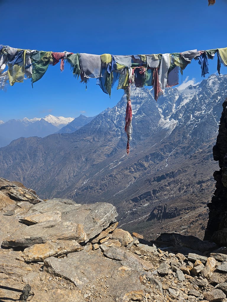 Himalayalar'da çekilmiş, renkli dua bandı ile çevrili dağ manzarası. Ağaçsız, kayalık bir alan ön planda, arka planda ise yüksek dağlar ve kar örtüsü görünmekte.