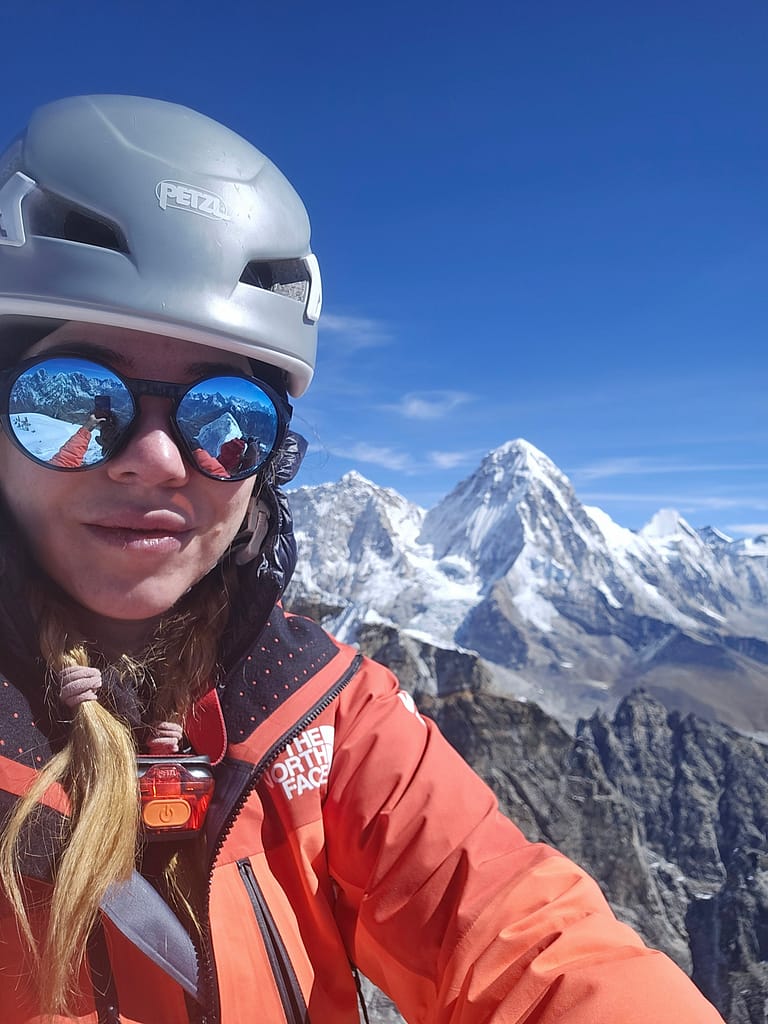 Bir dağcı, zirvenin arka planda göründüğü yüksek irtifada selfie çekiyor; kış montu ve kask takıyor, güneş gözlüğüyle çevresindeki dağ manzarasını izliyor.