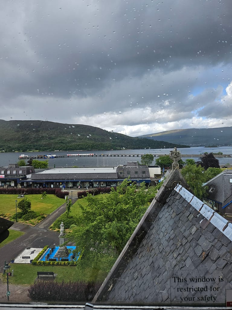 A landscape with gray clouds, green fields, and a fountain in the foreground. A lake and mountains are visible in the background. The view out the window is covered in water droplets.