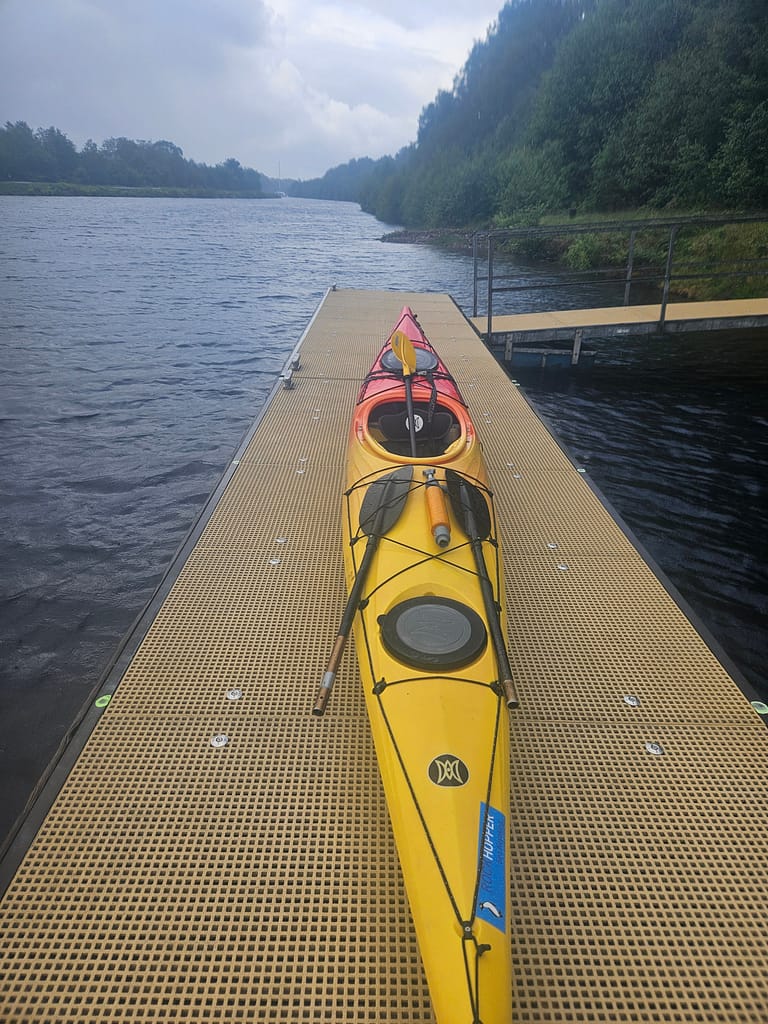 A yellow kayak sits on a dock, surrounded by a view of water and green woodland.