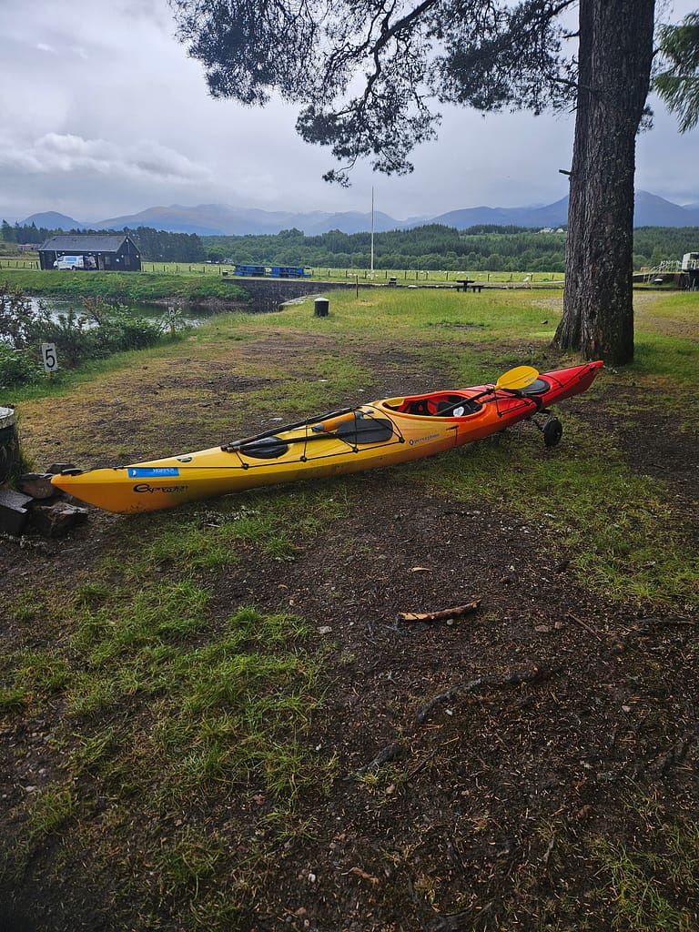 A yellow and orange ski stand in a field of green grass and trees. A mountain landscape can be seen in the distance. The weather is cloudy and rainy.