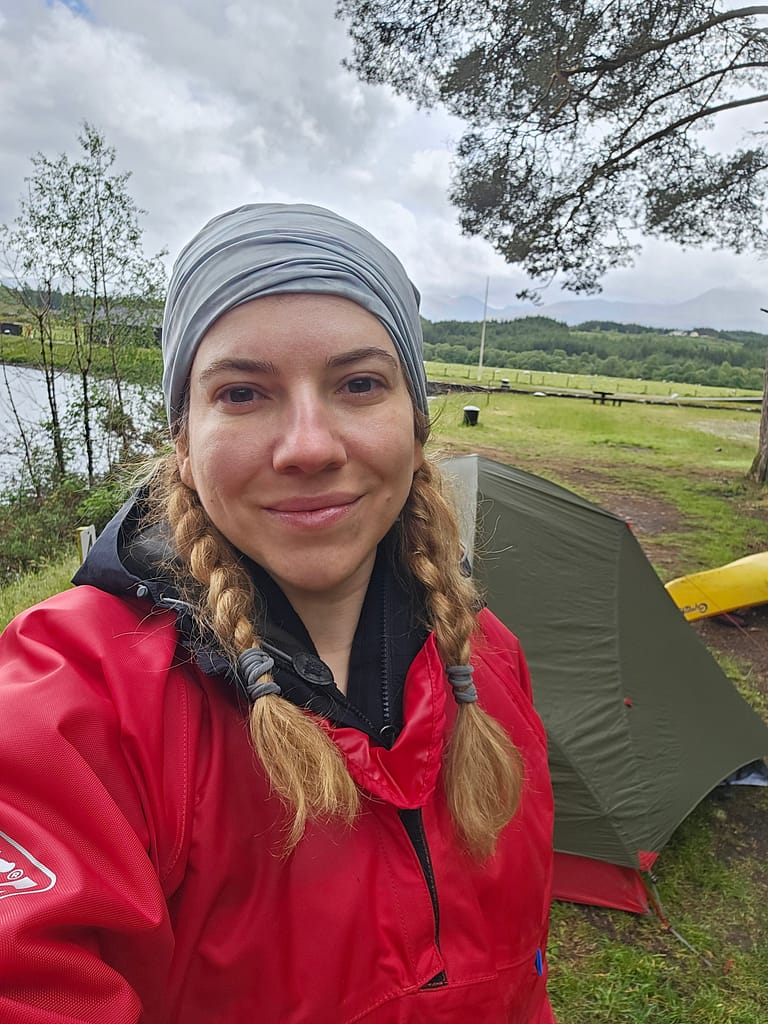 A woman takes a selfie in a red outerwear smiling with a tent and nature landscape in the background.