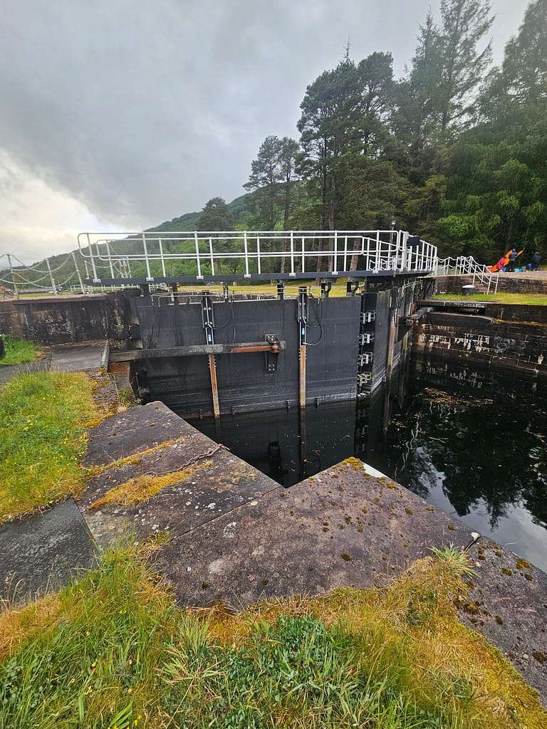 The image shows a water channel gate on the Great Glen Canoe Trail with surrounding greenery. The sky is cloudy, with trees visible in the background.