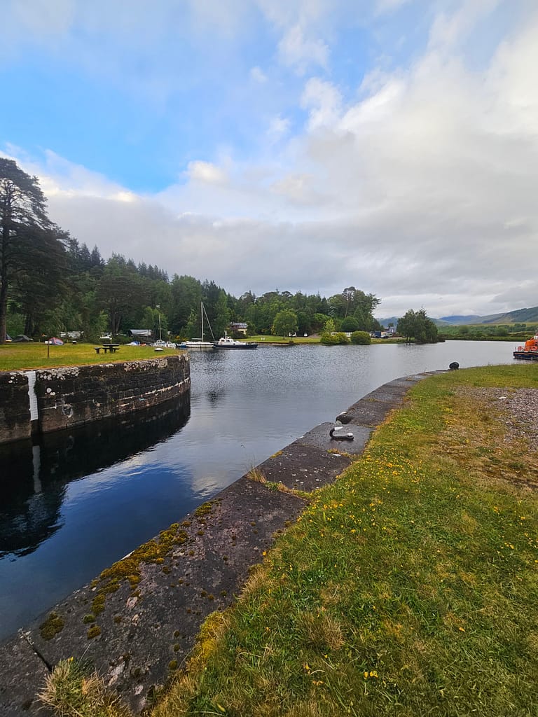 A waterway surrounded by stunning natural scenery. Green grass, trees, and a boat mooring line the shore. Reflections can be seen on the calm water.