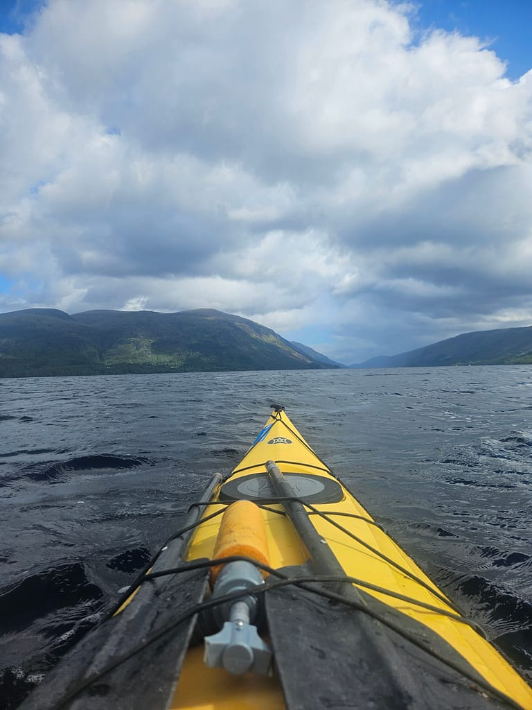 The view, taken from a canoe on the lake, shows clouds in the sky, gentle waves on the water, and mountainous areas in the background.