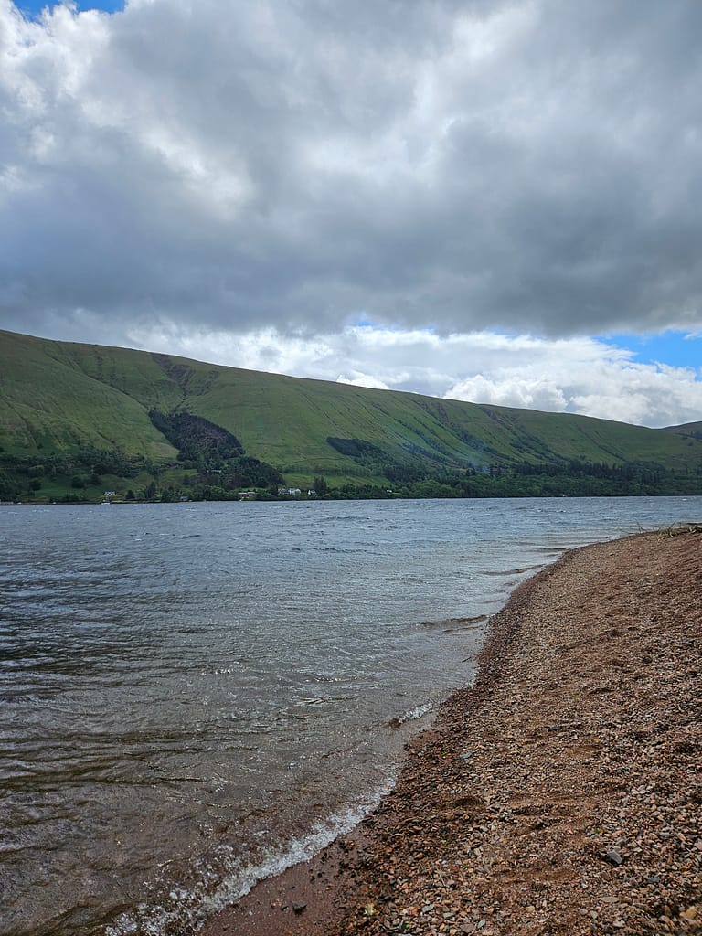 The image shows a lake surrounded by deep green slopes, a rocky beach along the lakeshore, and gray clouds dominating the sky.