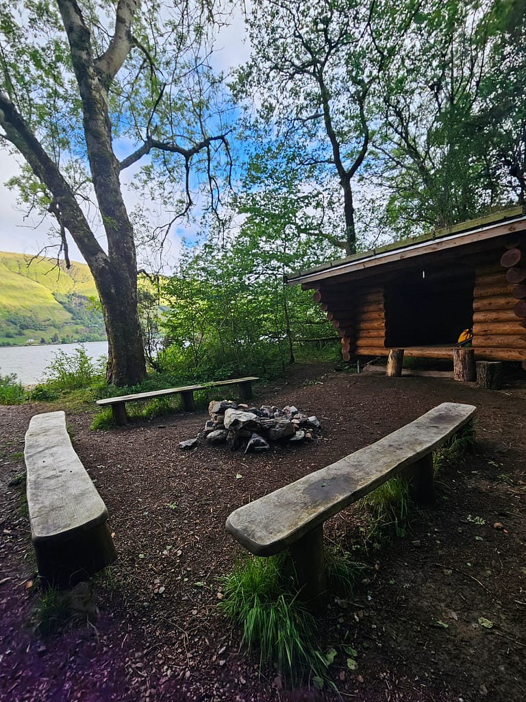 The campsite is in a forest area, surrounded by trees, with two long wooden chairs on the ground for sitting, and a fire pit in the middle.