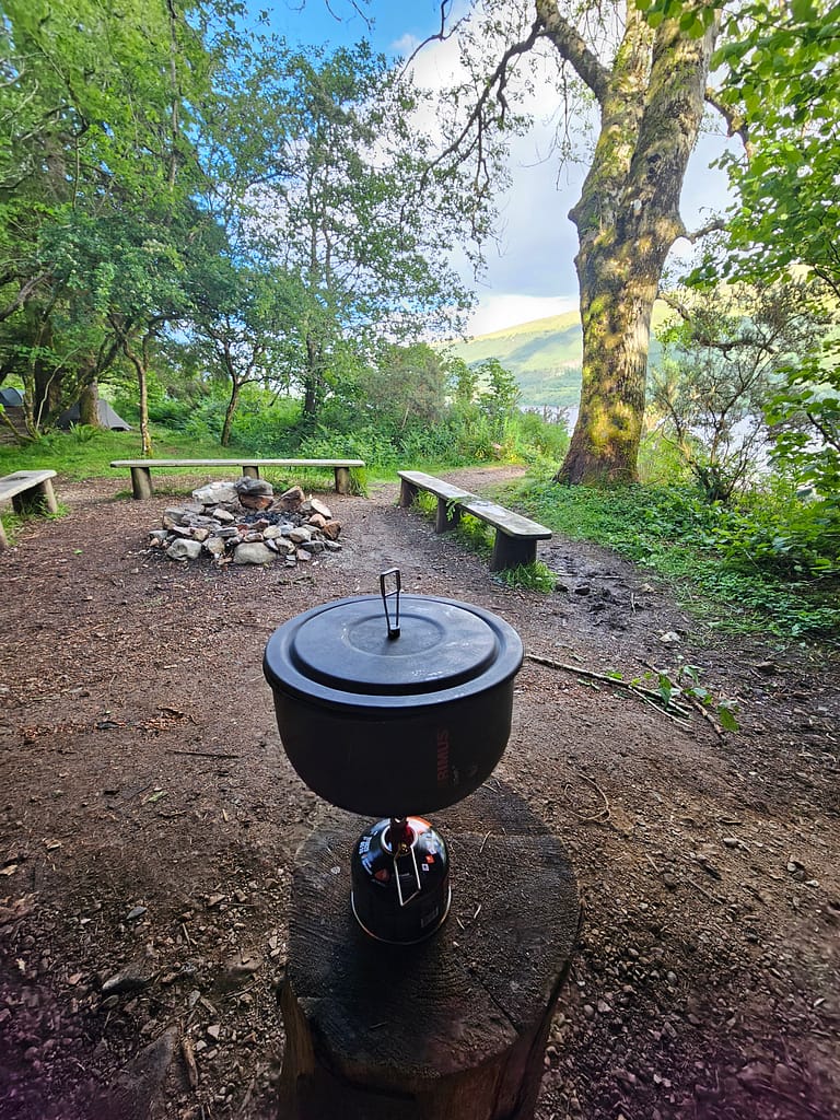 A campsite with a stove and wooden seating areas around it, lush green trees and a view of the sky.