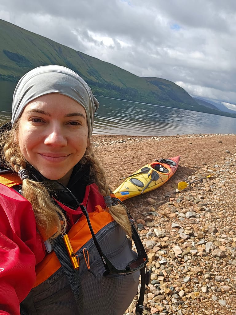 A woman smilingly takes a selfie by a lake in a canoe, with mountains and water visible in the background.