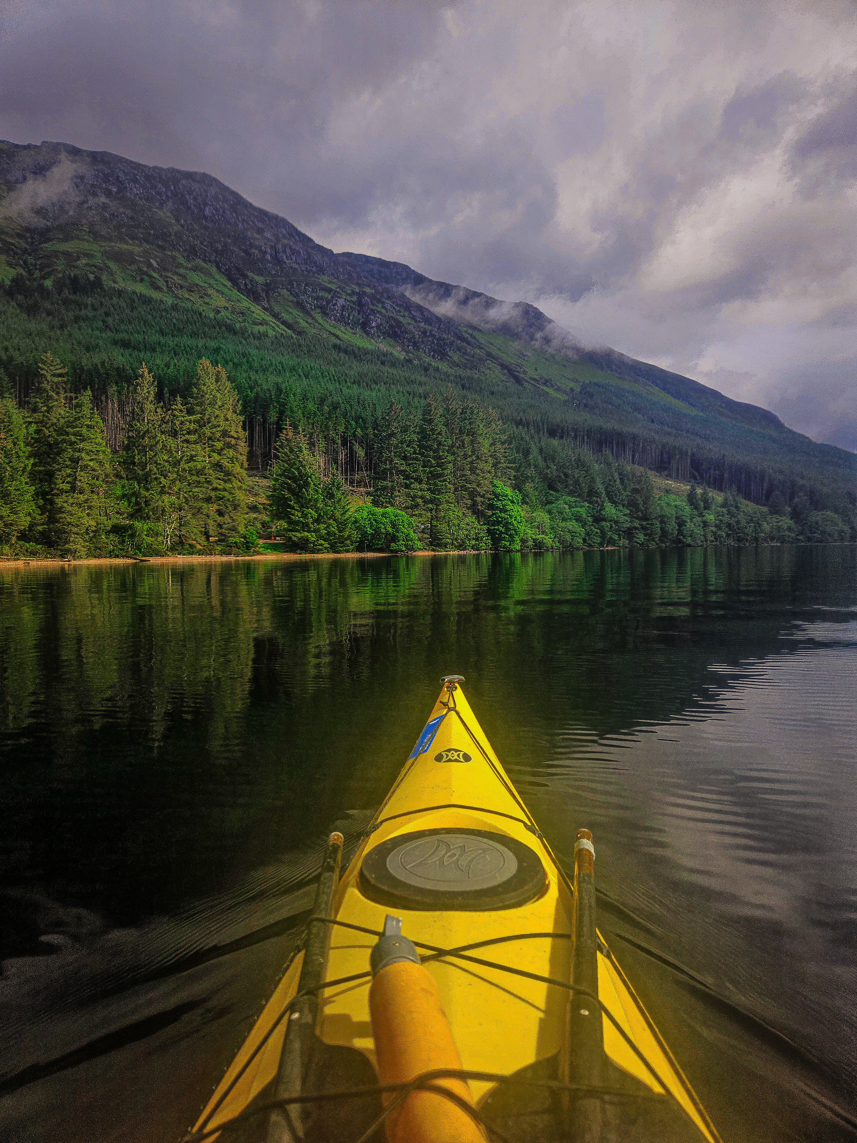 A person in a yellow kayak is cruising across a loch, surrounded by lush forests and mountain scenery. The sky is cloudy, and the water is calm.