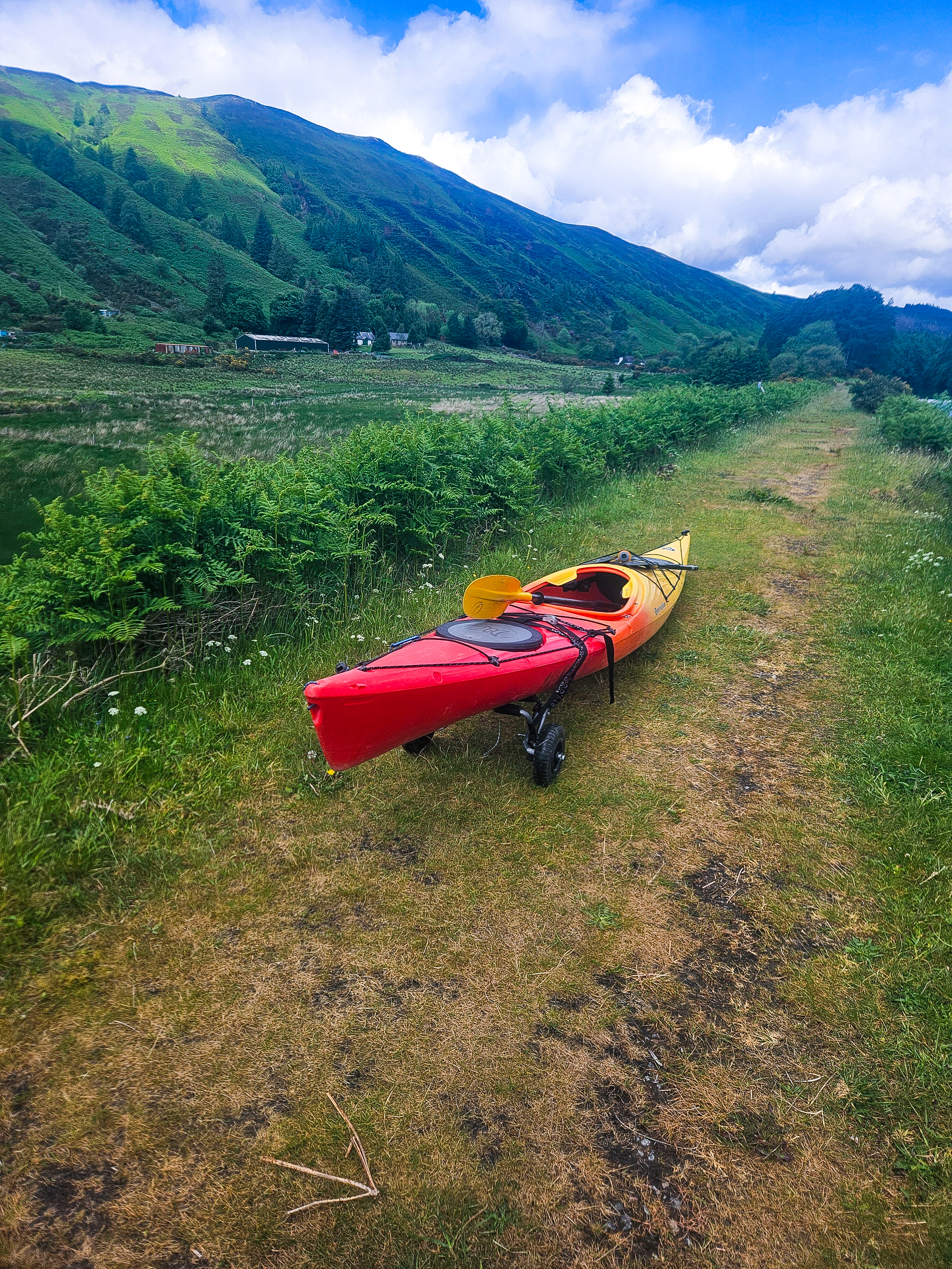 On a road, a canoe sits on a rock in the green grass. In the background, lush green mountains and a blue sky with white clouds can be seen.