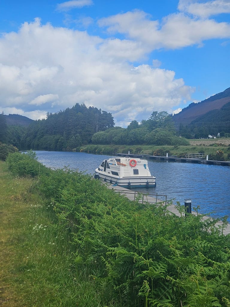 A beautiful natural scene; a boat parked on a waterway next to green fields with mountains and a cloudy sky in the background.