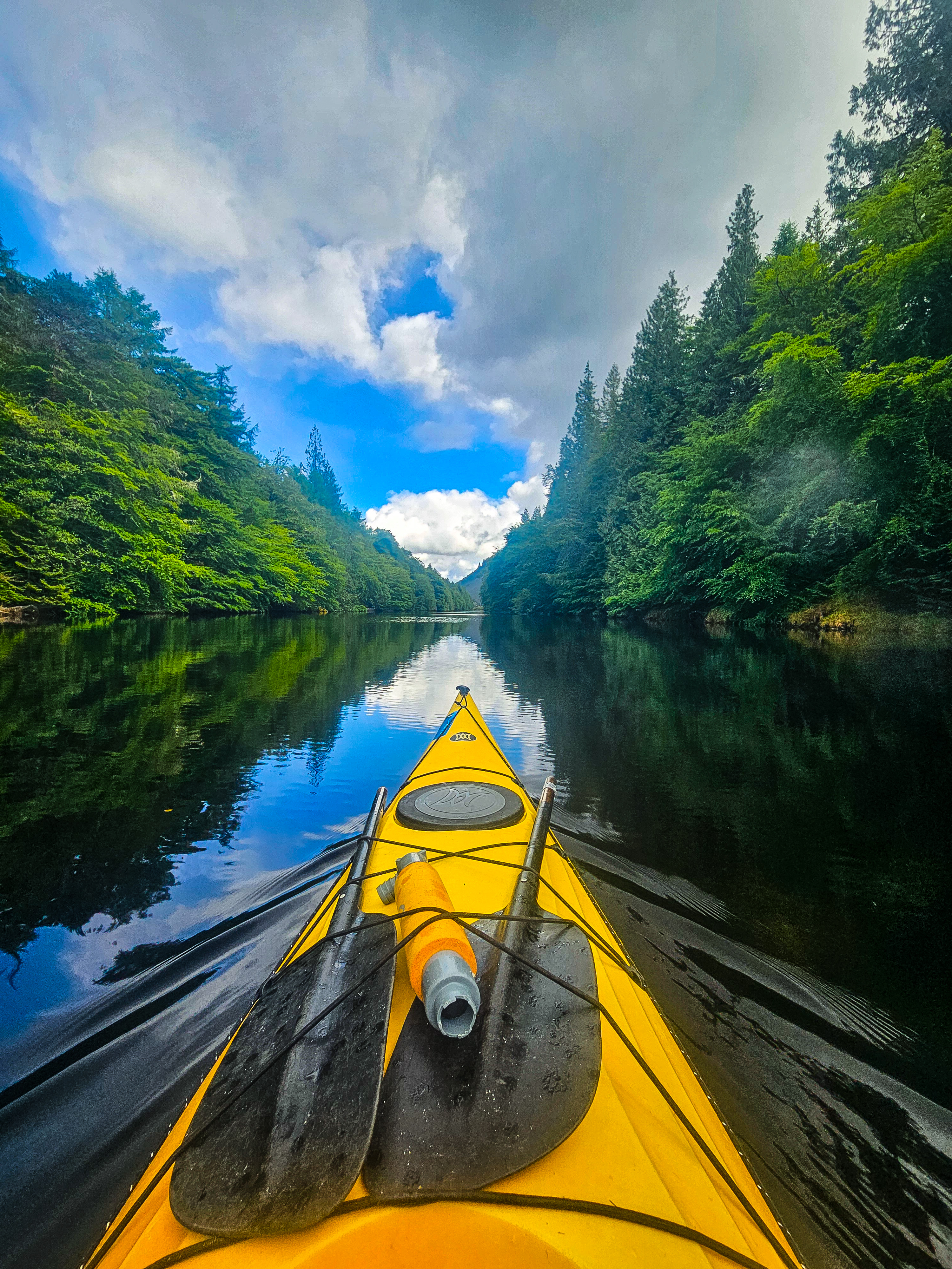 A yellow kayak navigates a peaceful waterway surrounded by trees. Clouds and blue holes in the sky create a beautiful reflection in the water.