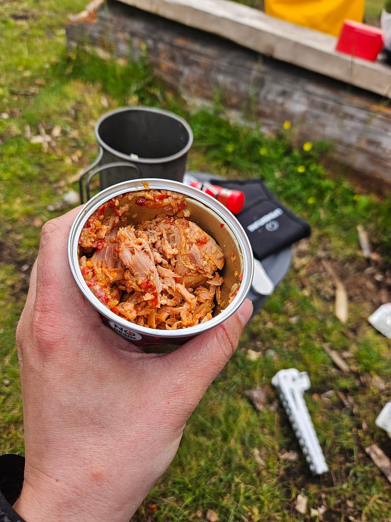 In one hand, an opened can of ground meat is held in place. In the background are camping equipment and a view of nature.