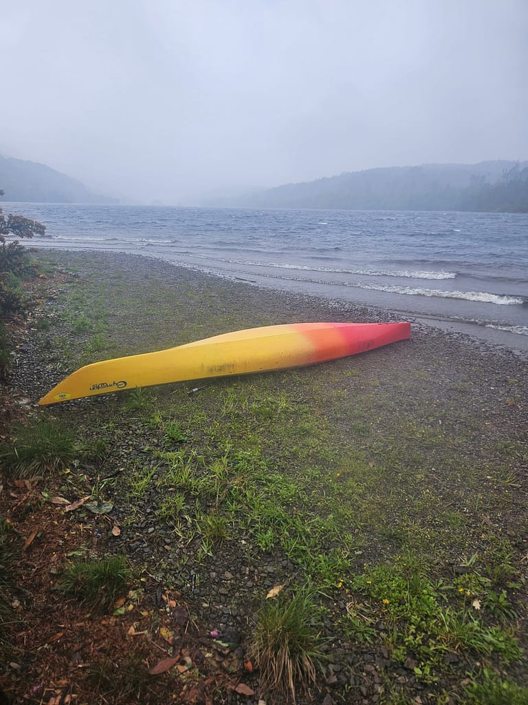 A yellow and orange canoe is seen abandoned at the edge of shallow water, with a rainy lakescape in the background.