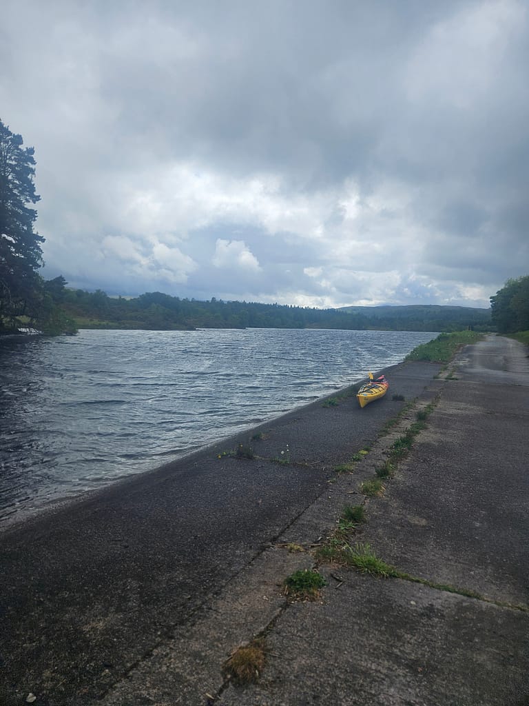 A yellow canoe on a shore, a serene water surface with gray clouds in the sky and a lush forest landscape.