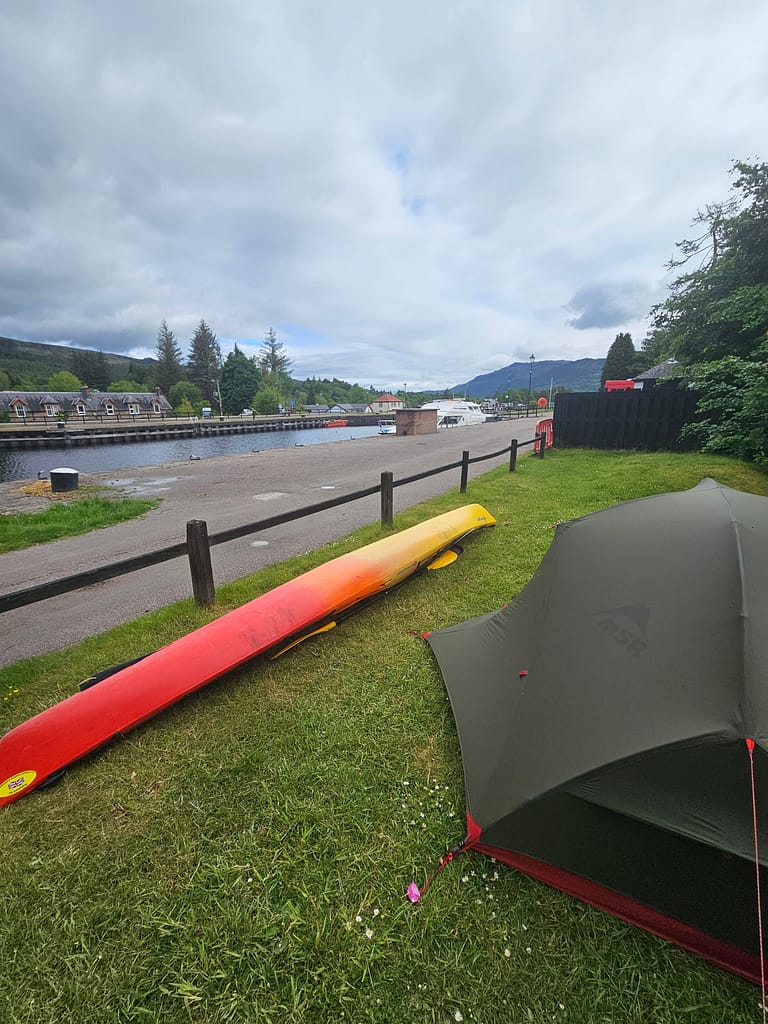 Camping site with a canoe and tent, located in a green area overlooking the canal.