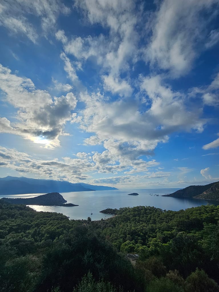 Fethiye'nin doğal güzelliklerini gösteren bir manzara, mavi gökyüzü ve bulutlarla dolu bir gün, deniz ve çevresindeki yeşil alanların görüntüsü.