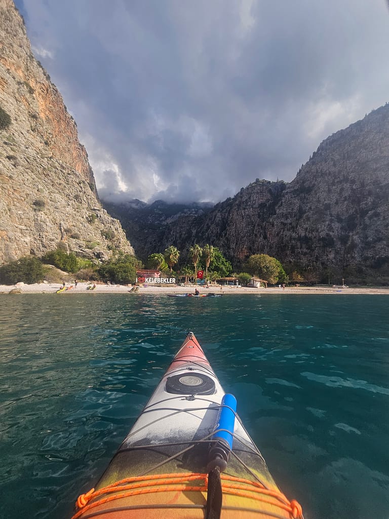 Bir deniz kayığı ile Kelebekler Koyu'nda ilerleyen bir kişinin perspektifi, arka planda yüksek kayalıklar ve yeşil bitki örtüsü görünüyor.