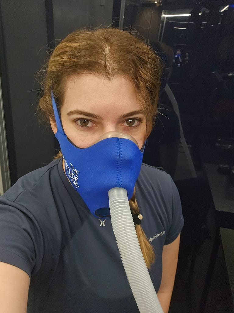 Bir kişi, yüksek irtifa simülatöründe maske takarak çekilmiş bir fotoğraf. Arka planda spor salonuna benzer bir iç mekan görünmekte.