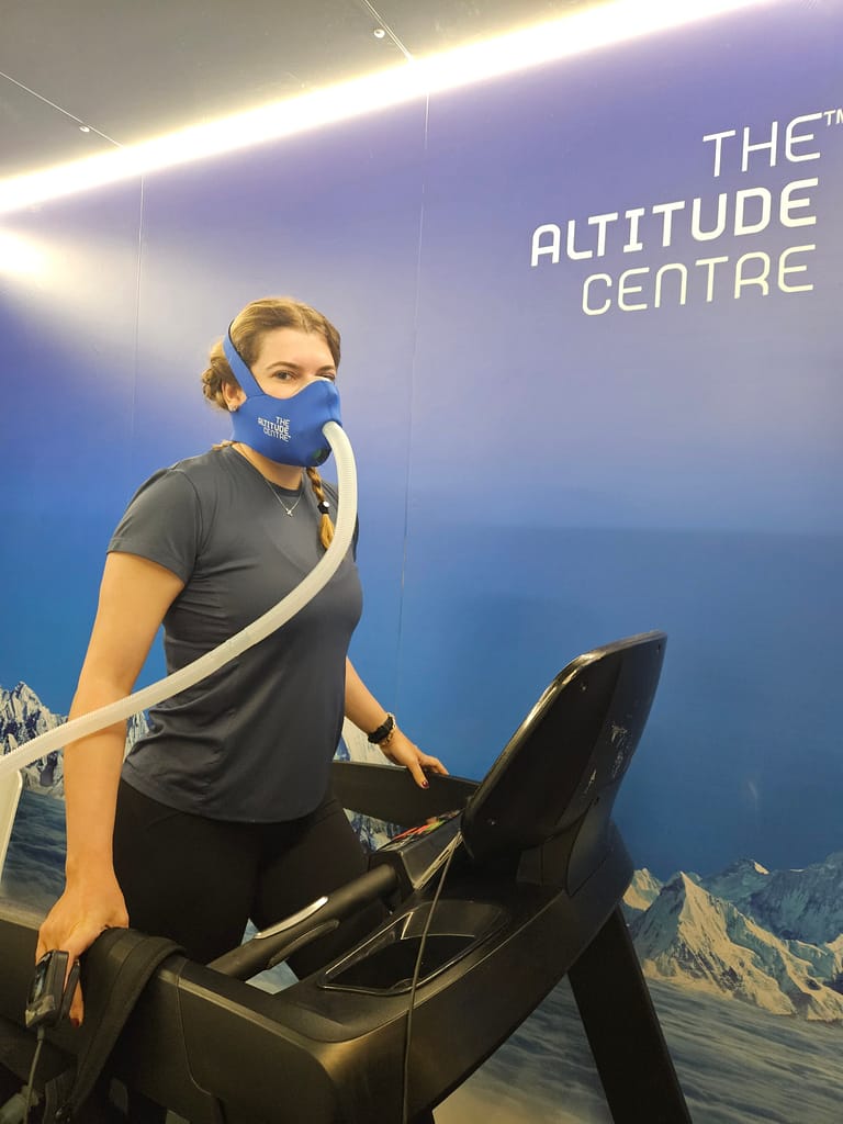 Bir kadın, yüksek irtifa simülatöründe koşu bandında antrenman yaparken oksijen maskesi takıyor. Arka planda, "THE ALTITUDE CENTRE" yazılı bir duvar görünmekte.