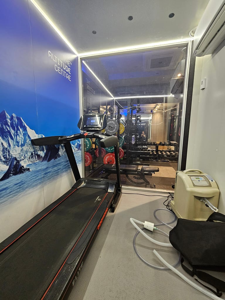 Bir yüksek irtifa simülatörünün iç görünümü, koşu bandı ve oksijen destek ünitesi ile birlikte. Arka planda ağırlık kaldırma ekipmanları görünmektedir.