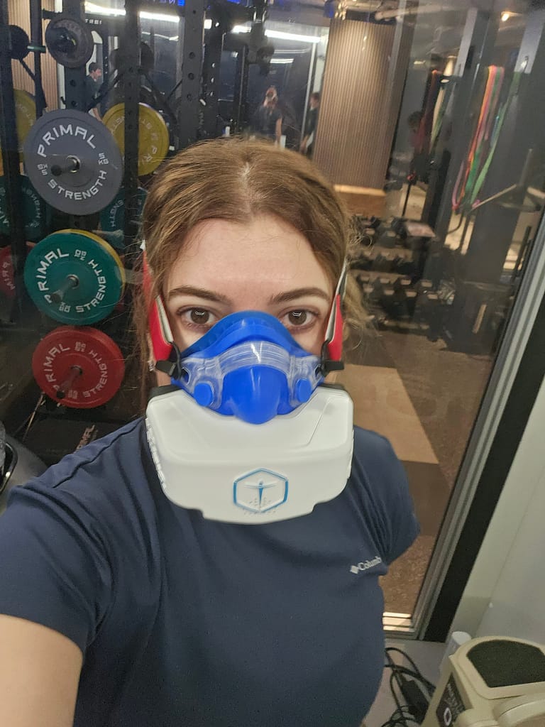 Bir spor salonunda yüksek irtifa simülatörü maskesi takan bir kişi, arka planda ağırlıklar ve spor ekipmanları ile görüntüleniyor.