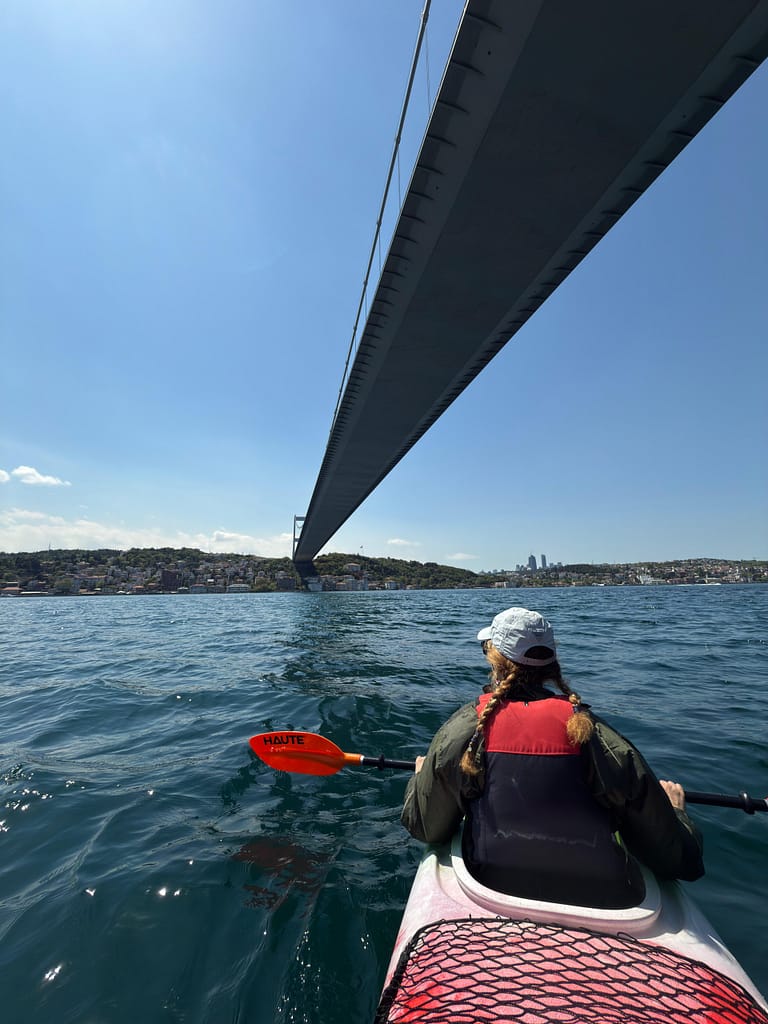 A person in a canoe is seen passing under a large bridge, with a city skyline and blue sky in the background.